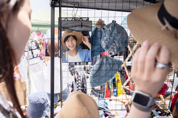 A young Asian woman and her friend happily try on hats in an outdoor clothing shop.
