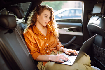 Business beautiful woman with a modern laptop sits in the back seat of a car. Successful startup, remote work. Freelance. 