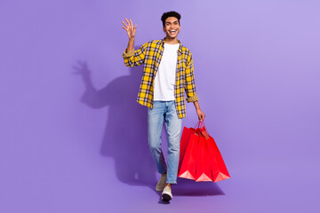Fototapeta premium Full body photo of funky cheerful person hold bags walking arm palm waving hi isolated on violet color background