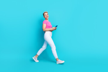 Fototapeta na wymiar Full size photo of positive cheerful woman dressed white trousers holding smartphone walk to empty space isolated on blue color background
