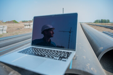 Laptop on pipes at a construction site. Reflection of a portrait of a builder in a helmet on the screen. 