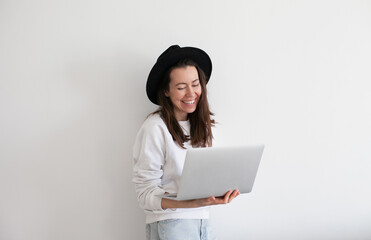 Young millennial woman wearing black hat using laptop, working online, white background. Copy space left and right.