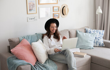 Young woman using laptop, working online, wearing black hat, smiling, sitting on a comfy couch. Trendy hipster cozy interior. Artist living slow freelance life. Millennial lifestyle. 