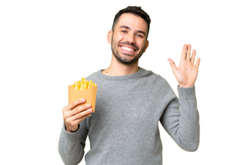 Young caucasian man holding fried chips over isolated chroma key background saluting with hand with happy expression