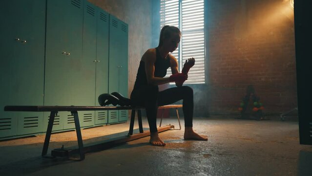 Serious woman fighter boxer wraps hands wrists with boxing bandage sits on bench in locker room, bottom view. Girl kickboxer prepares for fight. Professional sport, competition, preparation concept.