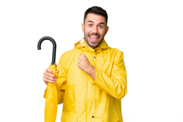 Young caucasian man with rainproof coat and umbrella over isolated background celebrating a victory