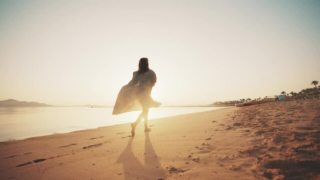 Carefree slim barefooted woman tourist in airy leopard pareo running along ocean beach leaving footprints on sand at sunset, back view. Girl stretching arms in sides feeling freedom on vacations.