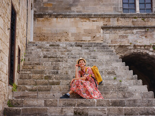 Young Asian woman in red dress walks through streets of Rhodes. woman resting on old house steps in fortress Rhodes. Tourism, vacation, and discovery concept, female traveler visiting southern Europe.