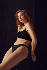 Fototapeta premium Portrait of fit woman in black underwear sitting on black chair and looking at camera on black studio background.