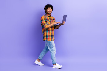 Full size photo of positive clever man wear checkered shirt jeans shoes hold laptop go empty space isolated on purple color background