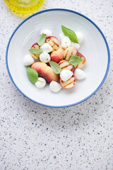 Salad with grilled peaches, mini mozzarella and basil, high angle view on a light-grey granite background, vertical shot with space