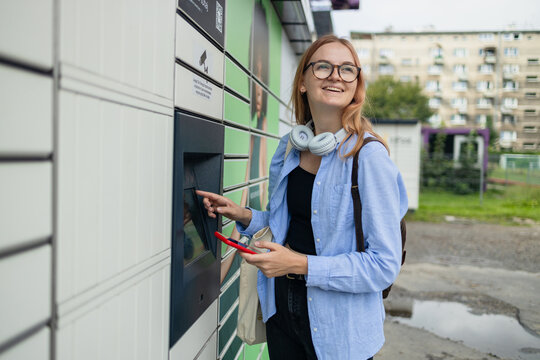 Beautiful woman picking up a package from a smart electronic steel parcel locker box, automatic mailboxes. Pachkomat delivery service, collection machine