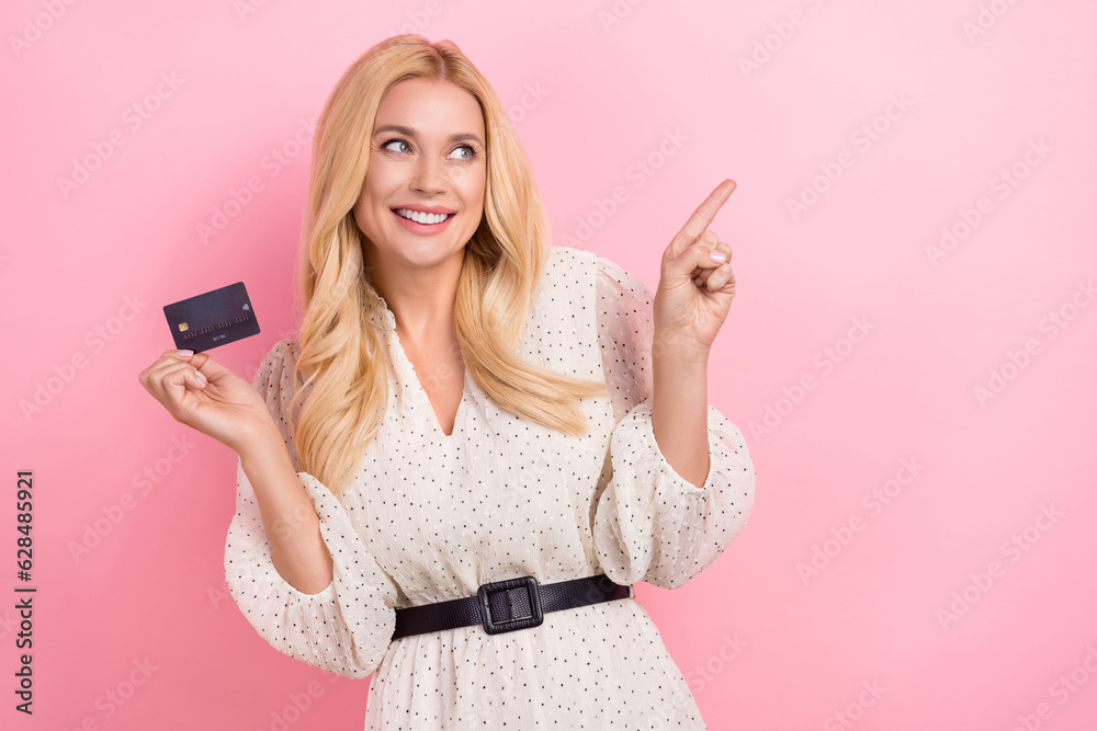 Wall mural Photo portrait of attractive blonde woman credit card finger point empty space wear trendy white clothes isolated on pink color background