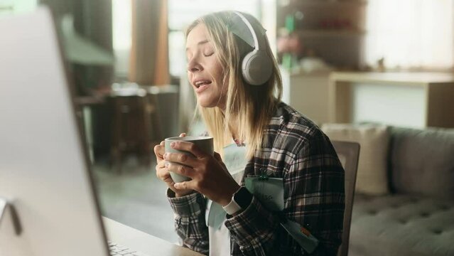 Cheerful blond mature woman freelancer with stickers listening to music in headphones drinking hot coffee or tea ignore deadline at distance remote work on computer at home No stress concept