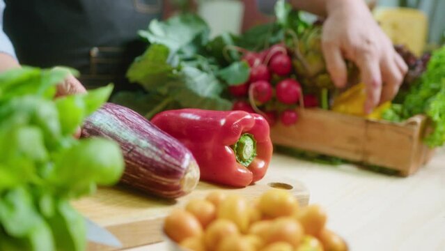 Male chef bringing wooden crate full of assorted fresh vegetables and placing eggplant, artichoke and bell peppers on kitchen table before cooking. Close-up shot