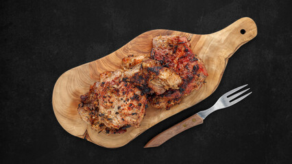 tasty grilled pork in natural olive wood cutting board beside the fork on dark tone texture background, top view, flat lay