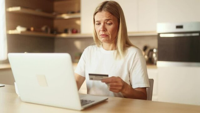 Confused blond woman with credit card thoughtfully pondering on screen while has problem with online order payment error or blocked bank account doing shopping at internet on laptop at light home
