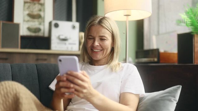 Portrait of charming blond mature woman hold smartphone scrolling watching social media indoors Happy female texting on her phone enjoying leisure time at home