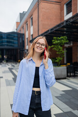 Fototapeta premium Vertical shot of happy young blonde woman makes telephone call in roaming wears casual clothes had glad expression walks against blurred city building enjoys smartphone talking. High quality photo