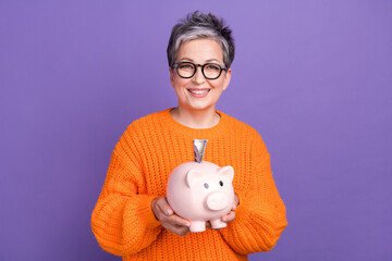 Photo of positive intelligent woman with short haircut dressed orange sweater hold pig money box isolated on purple color background