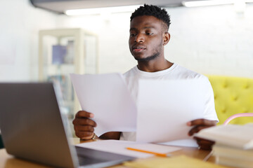 Concentrated black man who is sitting at a desk with a laptop and papers in front of them. They are a young and smart businessman, manager or entrepreneur who is working, thinking or doing finance.