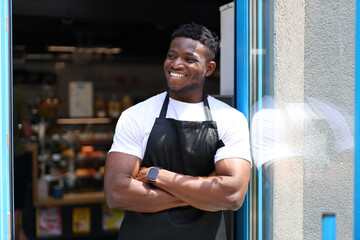 Successful black man who is wearing an apron and standing in front of a door. He is a young and...