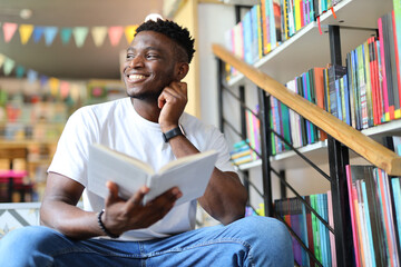 Happy and handsome black man sitting in a library or bookstore, reading a book. He is a young and...