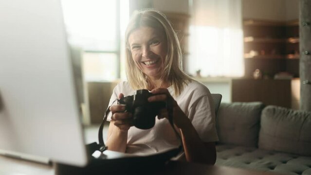 Portrait of pretty mature woman photographer hold digital camera looking at screen choosing photos for editing and looking at camera while sitting in front of computer at home workplace