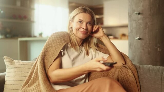 Portrait of happy positive mature blond woman with remote controller switches channels on TV and watching comedy movie show serial enjoying leisure time at home alone Fun weekend time