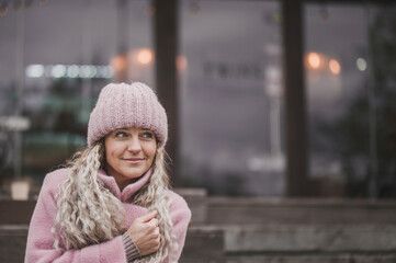 Fashionable mature woman in a pink knitted beanie hat and stylish coat outdoors in a cafe in autumn. Middle aged woman positive and smiling with pigtails in the city. Blur and selective focus