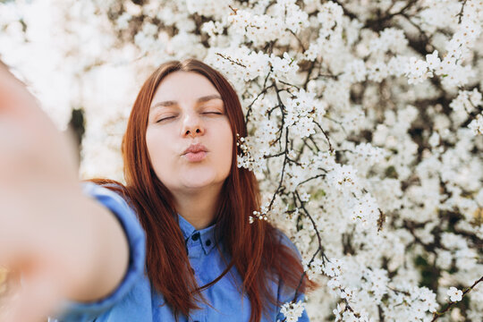 Young happy woman is making selfie on a camera on city street. Girl talking on a video call with phone outdoor. White flowers in the tree in the early spring. Flowers of the cherry blossoms