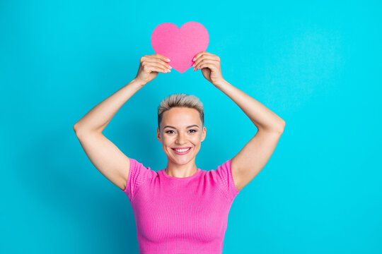 Photo of cheerful good mood pretty girl dressed pink t-shirt arms holding pink paper heart over head isolated on blue color background