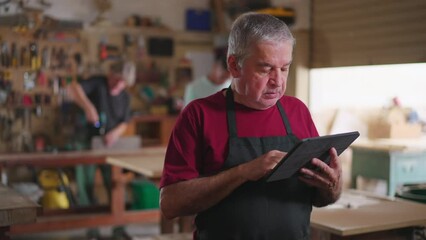 Senior worker wearing apron standing inside carpenter workshop holding tablet and browsing orders, job occupation concept using modern technology