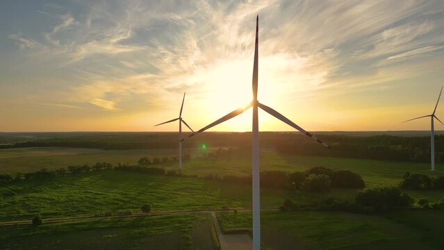 Large wind turbines with blades in field aerial view bright orange sunset blue sky wind park slow motion drone turn. Silhouettes windmills, large orange sun disc summer lens flare. Alternative energy