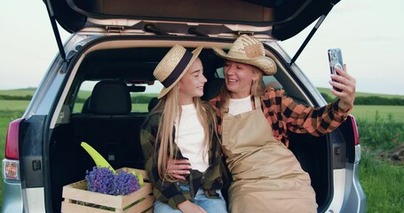 Family of farmers, mother and daughter dressed in aprons and hats sit in the trunk of a car next to a wooden box with fresh lavender taking selfie using phone in the field at sunset. Small family - Powered by Adobe