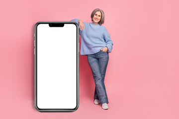 Full body length cadre photo of smile positive old woman wear blue knit jumper stay near big phone display menu isolated on pink color background
