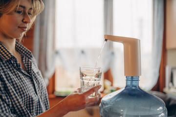 Woman's hands pour water into glass from an automatic water cooler, closeup top view. Pouring fresh clear water from blue huge bottle to glass. Clean drinking bottled water from the well.
