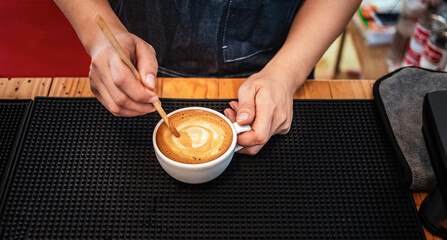 Close-up of the hand of a barista with a wooden spoon making or preparing coffee foam in a cup of...