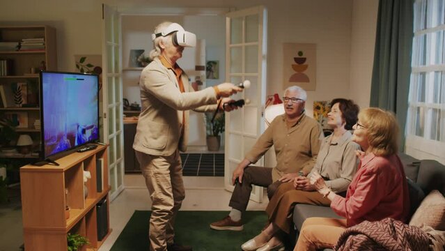 Long shot of company of elderly people looking at their friend using VR-glasses when hanging out together at home party