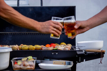 men hold beer in their hands on the background of the grill, vegetables and meat. Selective focus.