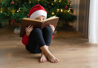 A little boy in a Santa Claus hat sits on the floor and hides behind a book with fairy tales