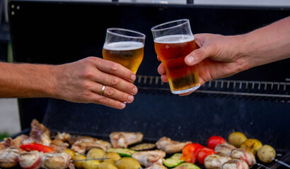 men hold beer in their hands on the background of the grill, vegetables and meat. Selective focus.
