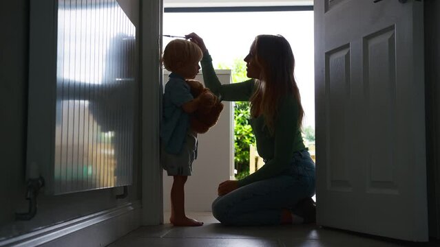 Mother measuring height of young son at home standing against wall in kitchen silhouetted against bright window - shot in slow motion