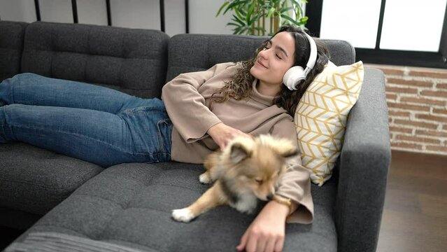 Young hispanic woman with dog listening to music lying on sofa at home