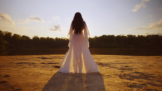 Happy woman walks along beach river banks on sand enters the water. Girl back rear view enjoy nature free green tree magic sun light sky. Lady walks in silk white long dress fly in wind slow motion
