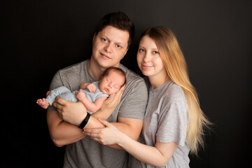 A cute newborn baby boy sleeps in a blue overalls in the first days of life. In the arms of father and mother. Warm parental hugs. On a black background. Professional portrait photography.