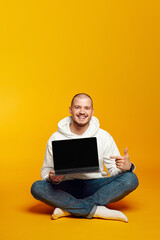 Vertical photo of hipster man wearing white hoodie smiling and pointing at new laptop in hands while sitting on floor, isolated over yellow background