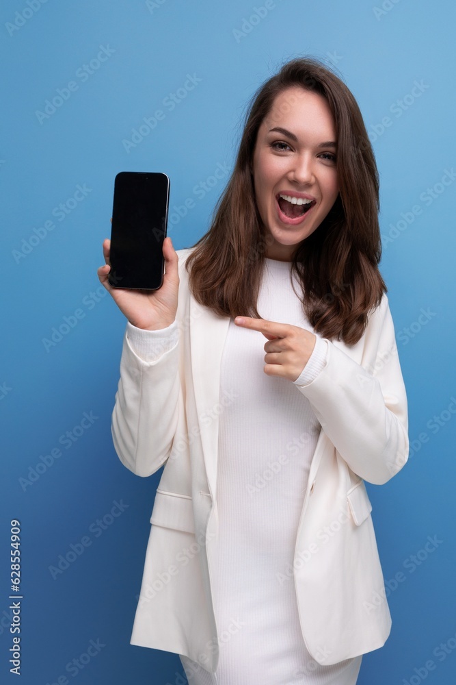 Wall mural smiling young brunette with long hair woman in white dress showing smartphone screen with mockup