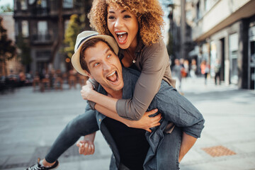 Fototapeta premium Young man carrying his girlfriend on a city street while they are traveling on their vacation