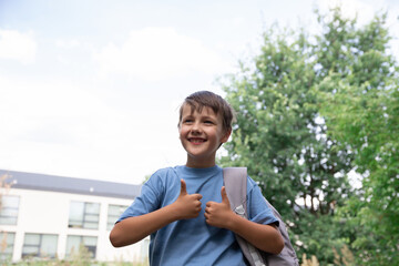 graduation or back to school. a cheerful little boy stands in front of the school. a child in a blue T-shirt and a gray backpack. the boy goes to school through the park.